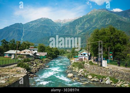 Ein kristallklarer Fluss schlängelt sich durch ein malerisches Dorf mit traditionellen Häusern, umgeben von lebhaften grünen Bergen unter einem hellblauen Himmel. Idee Stockfoto