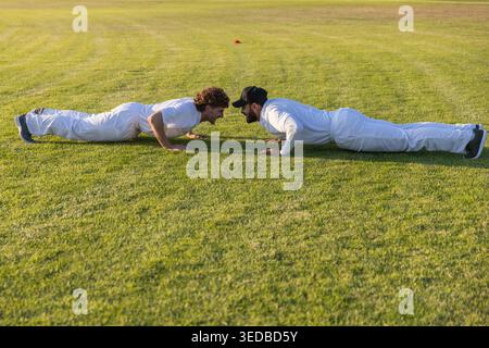 Zwei Männer, die Liegestütze auf dem Grasfeld in weißen Uniformen mit orangefarbenem Trainingskegel in der Nähe ausführen Stockfoto