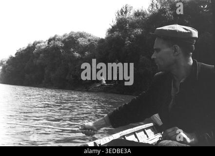 Donezk, 1960er Jahre, am offenen Fluss. Eine Profilaufnahme eines lokalen Fischers in flacher Mütze und dunkler Jacke, der in seinem Holzboot sitzt und eine Bambusrute hält. Dieser breitere Winkel zeigt die Weite des plätschernden Wassers und die ferne Küste, die von einem dichten, dunklen Wald unter einem hellen Sommerhimmel bedeckt ist. Dieses stimmungsvolle Schwarzweiß-Dokumentarfoto beleuchtet die einsame Romantik der ländlichen Amateurfischerei. Ein wunderschönes Vintage-Archiv, das perfekt das ruhige und friedliche Leben vor dem Krieg widerspiegelt. Stockfoto