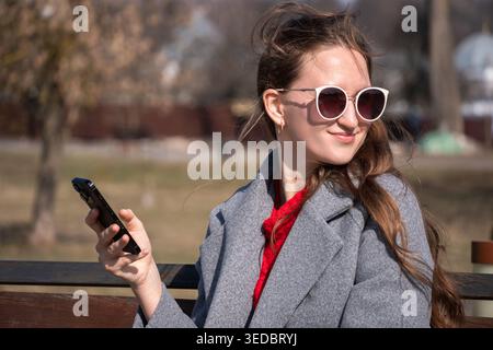 Eine junge Frau in rotem Pullover und Sonnenbrille sitzt auf einer Parkbank und lächelt, während sie an einem sonnigen Herbsttag ihr Smartphone überprüft und dabei eine sportart genießt Stockfoto