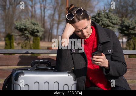 Frau, die auf einer Bank neben ihrem Koffer sitzt und Frustration und Stress erlebt, während sie ihr Smartphone nach Updates über eine Reiseverspätung oder einen Reiseanschlag sucht Stockfoto
