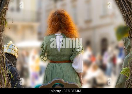 Rückansicht einer Frau mit leuchtend lockigen roten Haaren, die ein grünes Kleid im mittelalterlichen Stil trägt und bei einer Veranstaltung unter freiem Himmel inmitten einer verschwommenen Menschenmenge steht Stockfoto