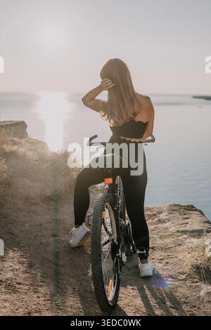 Frau Fahrrad Klippe Rückansicht einer jungen Frau auf ihrem Fahrrad mit Blick auf das Meer während Sonnenuntergang für ein aktives Lifestyle-Konzept Stockfoto