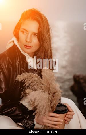 Frau Sunset Coffee Junge Frau mit Lederjacke sitzt am Meer bei Sonnenuntergang und hält Papierbecher und Pampas Gras Stockfoto