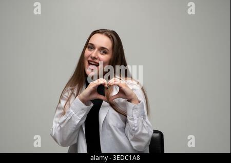 Eine junge Frau lächelt mit offenem Mund, zeigt mit ihren Händen eine Herzform, vermittelt Liebe, Sorgfalt und Glück Stockfoto