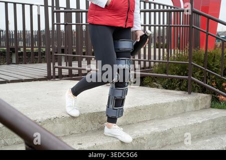 Frau mit Sportkleidung und Kniestraße oder Orthese nach einer Beinoperation, die Treppe im Park hinunter läuft. Medizin- und Gesundheitskonzept. Stockfoto