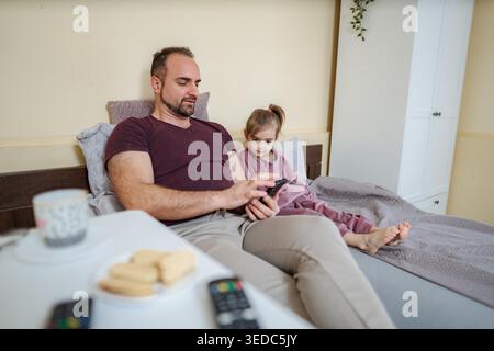 Vater und Tochter entspannen sich im Bett, während sie auf den Smartphone-Bildschirm schauen Stockfoto