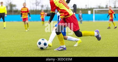 Jugendfußballspieler im roten und gelben Uniform Dribbling Ball während des Wettkampfs auf Green Grass Field - Junior Football League Action Stockfoto
