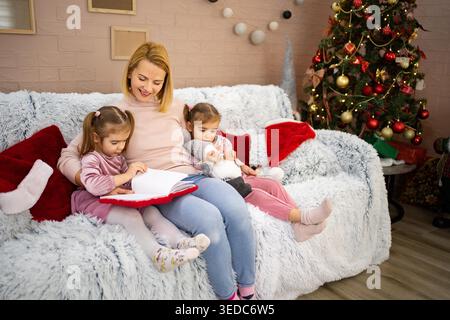 Weihnachten: Mutter liest ihren beiden Töchtern eine Geschichte vor, während sie auf dem Sofa neben dem Weihnachtsbaum sitzt Stockfoto