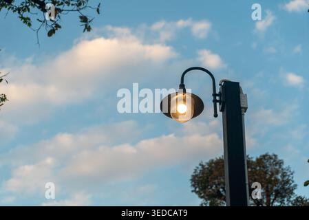 Eine wunderschöne Straßenlaterne im Freien, die einen warmen Glanz vor einem blauen Himmel mit flauschigen Wolken wirft und jedem städtischen Ambiente Charme und Beleuchtung verleiht. Stockfoto