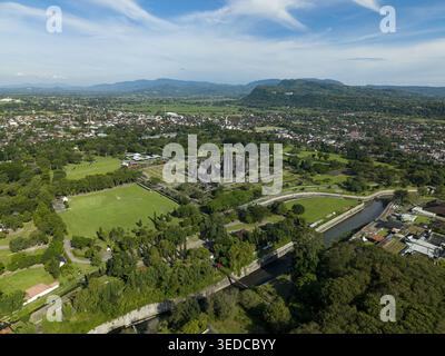 Blick aus der Vogelperspektive auf den Prambanan Tempel mit antiken Steintürmen, üppiger grüner Parklandschaft, einem gewundenen Fluss und fernen Bergen unter einem blauen Himmel in Pramba Stockfoto