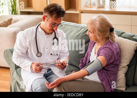 Ein medizinisches Fachpersonal kontrolliert den Blutdruck einer älteren Patientin, während sie zu Hause bequem auf einer Couch sitzt. Stockfoto