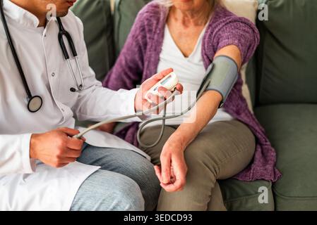 Nahaufnahme eines medizinischen Fachpersonals, das den Blutdruck eines älteren Patienten kontrolliert, während sie zu Hause bequem auf einer Couch sitzt. Stockfoto