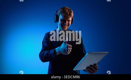 Ein lächelnder Mann, der die Daumen hoch hält, während er Tablet und Kopfhörer benutzt Stockfoto