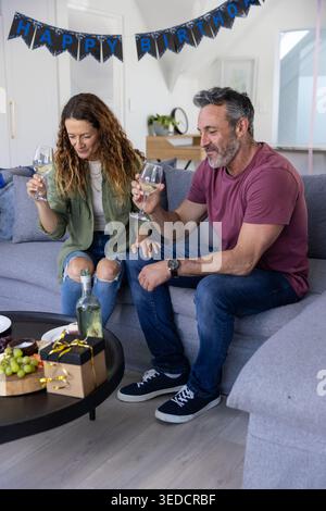 Seniorenpaar, das auf grauem Sofa sitzt und Weingläser unter HAPPY BIRTHDAY Banner hält, Geschenk auf dem Tisch Stockfoto