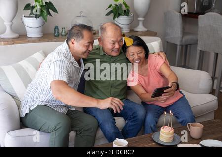 Asiatische Familie macht zu Hause Selfie auf Sofa mit Smartphone, Kuchen, Senioren mit pfirsichfarbenem T-Shirt Stockfoto