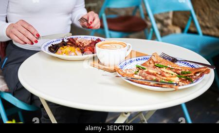 Frau genießt Brunch im Café im Freien mit herzhaftem Crepe, englischem Frühstück und Cappuccino, zwangloser Speiseszene, Lifestyle-Konzept, Wochenendentspannung, geselliges Essen Stockfoto
