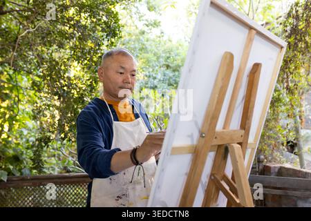 Ein älterer asiatischer Mann in bemalter Schürze, der auf einer Veranda steht und Leinwand auf einer Staffelei mit Pinsel hält Stockfoto