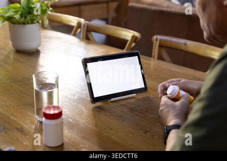 Senior Mann sitzt am Esstisch und hält eine Pille-Flasche, liest Etikett neben Tablette, Kopierraum Stockfoto