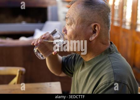 Ein älterer asiatischer Mann sitzt am hölzernen Esstisch mit klarem Glas und Trinkwasser, Kopierraum Stockfoto