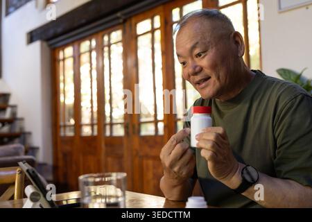 Leitender asiatischer Mann, der die Pille-Flasche mit roter Kappe am Holztisch mit Tablette hält und inspiziert Stockfoto