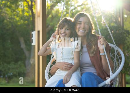 Weibliche Mutter und Tochter sitzen auf hängendem, gewebtem Sessel, schaukeln im Garten und lächeln Stockfoto