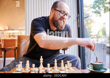 Ruhiger Mann, der Kaffee nach dem Spiel zubereitet. Ruhiger, individueller Kaffee in warmer Atmosphäre. Whitehaar-Mann rührt friedlich Kaffee neben Schachfiguren mit sanftem Lächeln Stockfoto