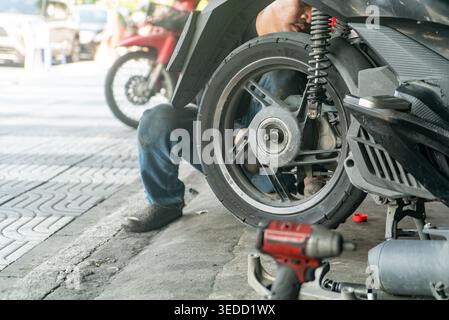 Ein Mechaniker, der in einer belebten Werkstatt im Freien einen Motorradreifen fleißig repariert. Werkzeuge und Teile sind verstreut und zeigen praktische Wartung Stockfoto