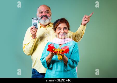 Ein älteres Seniorenpaar in Indien im Alter, das zusammen steht und Goldschmuck und Banknoten der Indischen Rupie hält, während er auf die Kamera isola zeigt Stockfoto