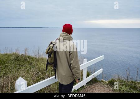 Reisender in roter Mütze und khakifarbener Jacke mit Rucksack, der auf einer Klippe steht und auf das Meer blickt Stockfoto