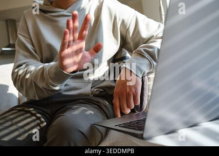 Mann in einem beigefarbenen Hoodie hält eine Hand in einer „Stop“-Geste an einen Laptop mit Sonnenlicht- und Schattenmuster Stockfoto
