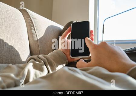 Mann sitzt auf einem Sofa und hält ein Smartphone mit einem leeren schwarzen Bildschirm in einem sonnendurchfluteten Raum Stockfoto