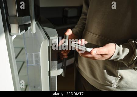 Mann in einem beigefarbenen Hoodie, der ein Smartphone hält, während er einen modernen silbernen Kühlschrank in einer sonnenbeleuchteten Küche öffnet Stockfoto