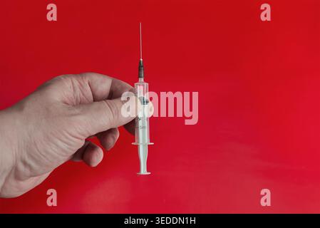 Nahaufnahme einer Hand, die eine medizinische Einwegspritze hält, mit der Nadel nach oben auf einem hellroten Hintergrund, Konzept der Impfung, Nothilfe, Stockfoto