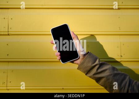 Hand hält ein Smartphone mit einem leeren schwarzen Bildschirm vor einem leuchtend gelben Holzhintergrund bei hellem Tageslicht Stockfoto