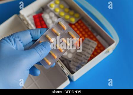 Hand in einem blauen medizinischen Handschuh, der eine Blisterpackung mit bunten Pillen vor dem Hintergrund eines offenen erste-Hilfe-Kits mit verschiedenen bunten Tabletten hält, Clos Stockfoto