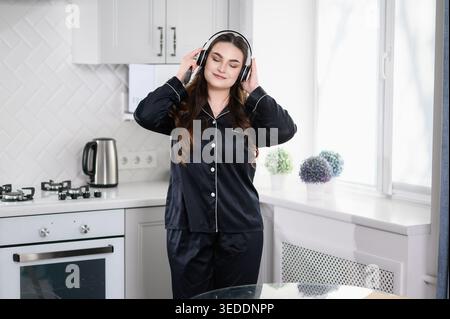 Eine ruhige junge Frau im schwarzen Seidenschlafanzug steht in der hellen Küche und hört Musik mit geschlossenen Augen. Erfasst Konzepte der psychischen Gesundheit, Stress rel Stockfoto