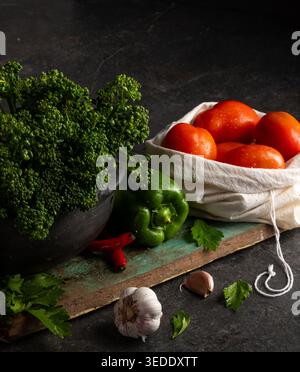 Frisches Obst und Gemüse steht auf einem Holzbrett in einer Küche. Grüne Kräuter, rote Tomaten und Knoblauch werden mit Paprika und Blattgemüse serviert. Natu Stockfoto