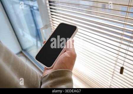 Person, die ein Smartphone mit einem leeren schwarzen Bildschirm in der Nähe eines Fensters mit Holzjalousien hält Stockfoto