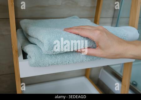 Die Hand der Person legt oder berührt einen Stapel weicher hellblauer Handtücher auf einem weißen Holzregal im Badezimmer Stockfoto