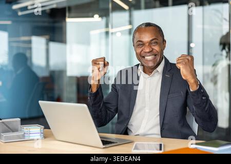 Ein erwachsener Unternehmer sitzt an seinem Schreibtisch, weht fröhlich die Fäuste in einer Geste der Leistung und des Glücks und arbeitet mit einem Laptop in einer hellen Unternehmensumgebung Stockfoto