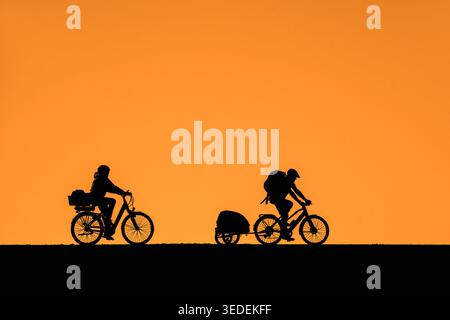Das junge Paar mit Fahrradhelmen, die Frau, die auf dem E-Bike fährt, und der Mann zieht den Fahrradanhänger, die Silhouette vor dem orangefarbenen Sonnenuntergangshimmel im Sommer Stockfoto