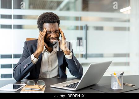 Geschäftsmann, der in einem Büro die Bügel umklammert, die Augen an Laptop und Tablet geschlossen, Migräne, Stress und Burnout durch lange Stunden, Müdigkeit und überwältigenden Arbeitsdruck zeigt Stockfoto