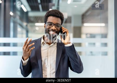 Geschäftsmann in Anzug und Brille lächelt und bewegt während eines ansprechenden Mobiltelefongesprächs in einem modernen Büro, vermittelt eine sichere Kommunikation, Erfolg und professionelle Verbindung Stockfoto
