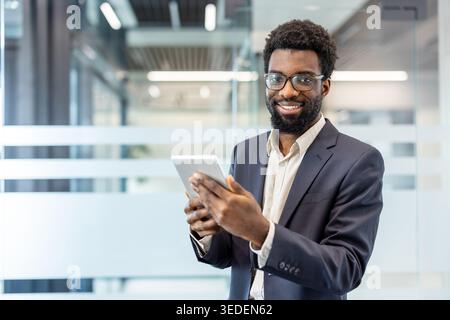 Junger, erwachsener Geschäftsmann, der in die Kamera lächelt, ein digitales Tablet hält und selbstbewusst in einem modernen Glasbüro steht und Technologie und Erfolg im Geschäft repräsentiert Stockfoto