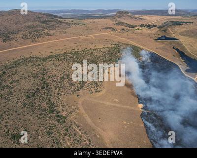 Aus der Vogelperspektive eines kontrollierten Buschfeuers mit dickem weißem Rauch, der aus verkohltem Grasland und sanften Hügeln unter einem klaren blauen Himmel in Waterberg Distri aufsteigt Stockfoto