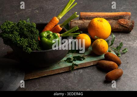 Frisches Obst und Gemüse wird in eine Schüssel auf dunkler Oberfläche gelegt. Grüner Brokkoli, Orangen-Zitrusfrüchte, Karotten und ein grüner Paprika sind sichtbar Stockfoto