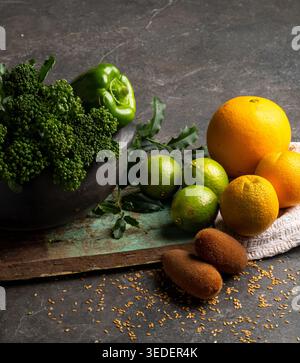 Eine Schüssel hält grünen Brokkoli und grüne Paprika neben Orangen, Limetten und ein paar anderen Früchten auf einem Holzbrett. Ein Tuch liegt unter den Früchten auf a d Stockfoto