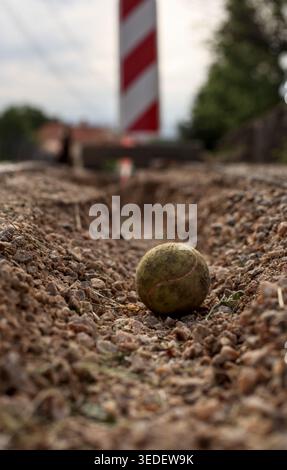 Ein alter Tennisball in einer Schmutzrille auf Kiesboden. Verschwommene rot-weiße Straßensperre im Hintergrund unter einem bewölkten Himmel. Stockfoto
