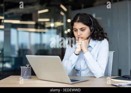 Gestresste junge Frau im Headset an einem modernen Bürotisch, starrte mit Erschöpfung und Frustration auf den Laptop, erlebte Burnout und Enttäuschung während der Fernarbeit Stockfoto